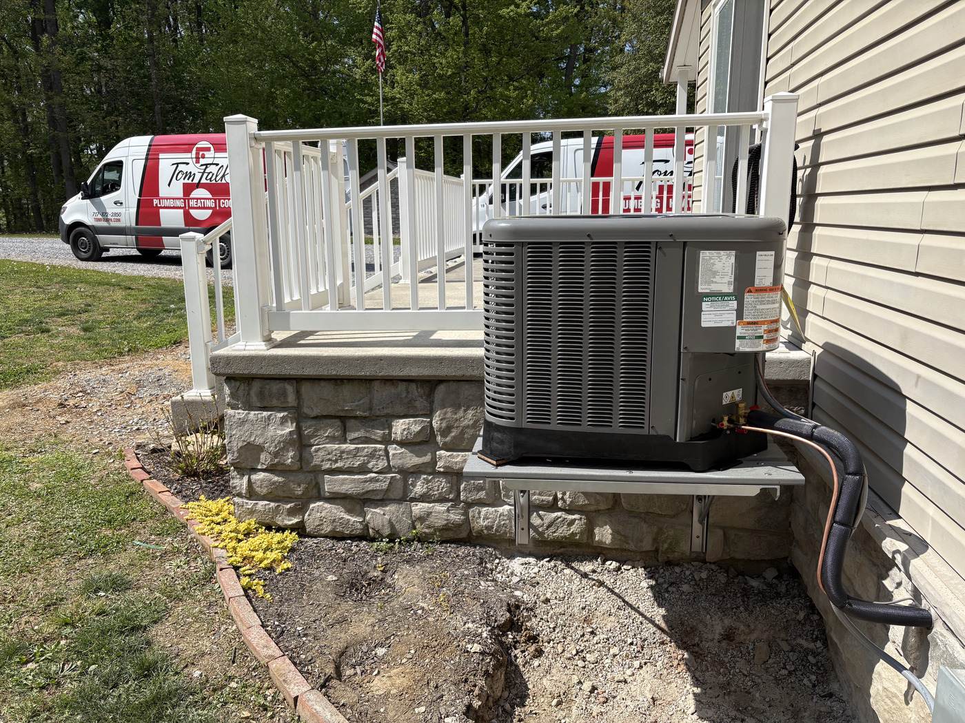 Two branded Tom Falk service vans and an American flag beside a newly installed air-conditioning condenser.