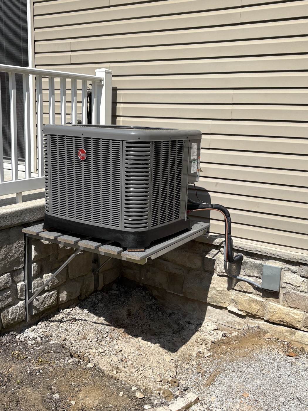 Rheem air-conditioning condenser on a wall bracket outside a home, installed by Tom Falk.