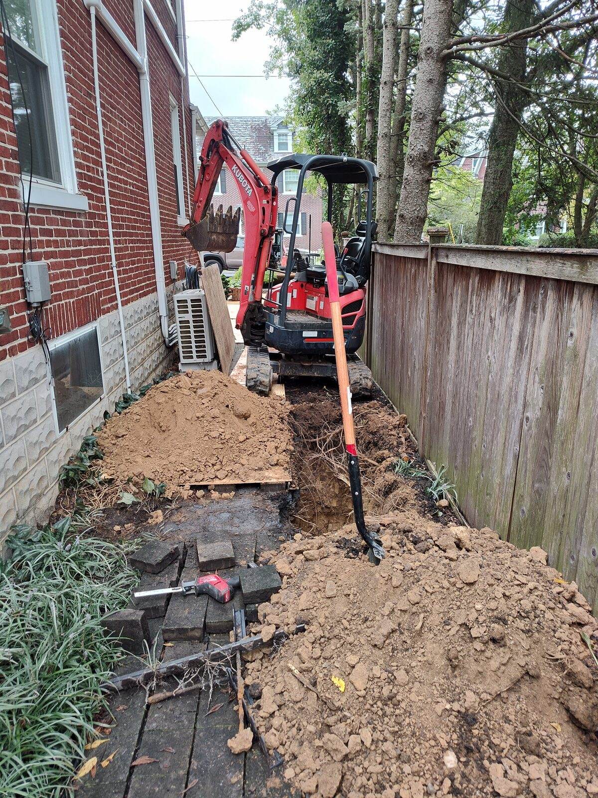 A compact red Kubota excavator digging a narrow trench in the tight space between a brick house and a wood privacy fence.