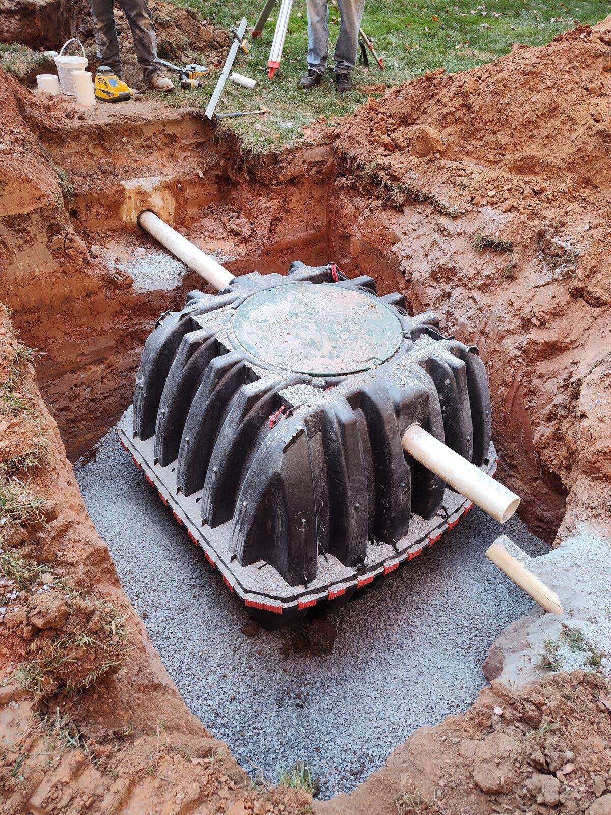 A new ribbed black poly septic tank set on a gravel bed in a fresh excavation, inlet and outlet pipes connected, on a Lancaster County property.